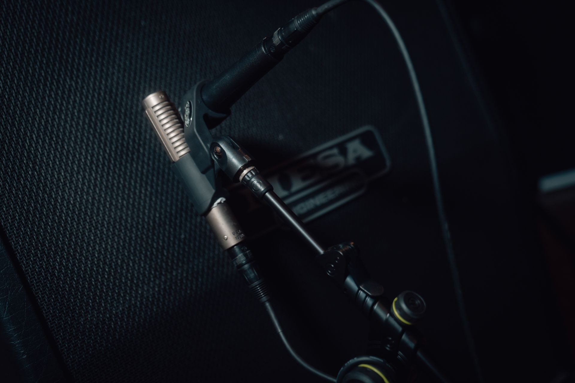 Close-up of a condenser microphone setup with a Mesa amplifier in the background under dim lighting.