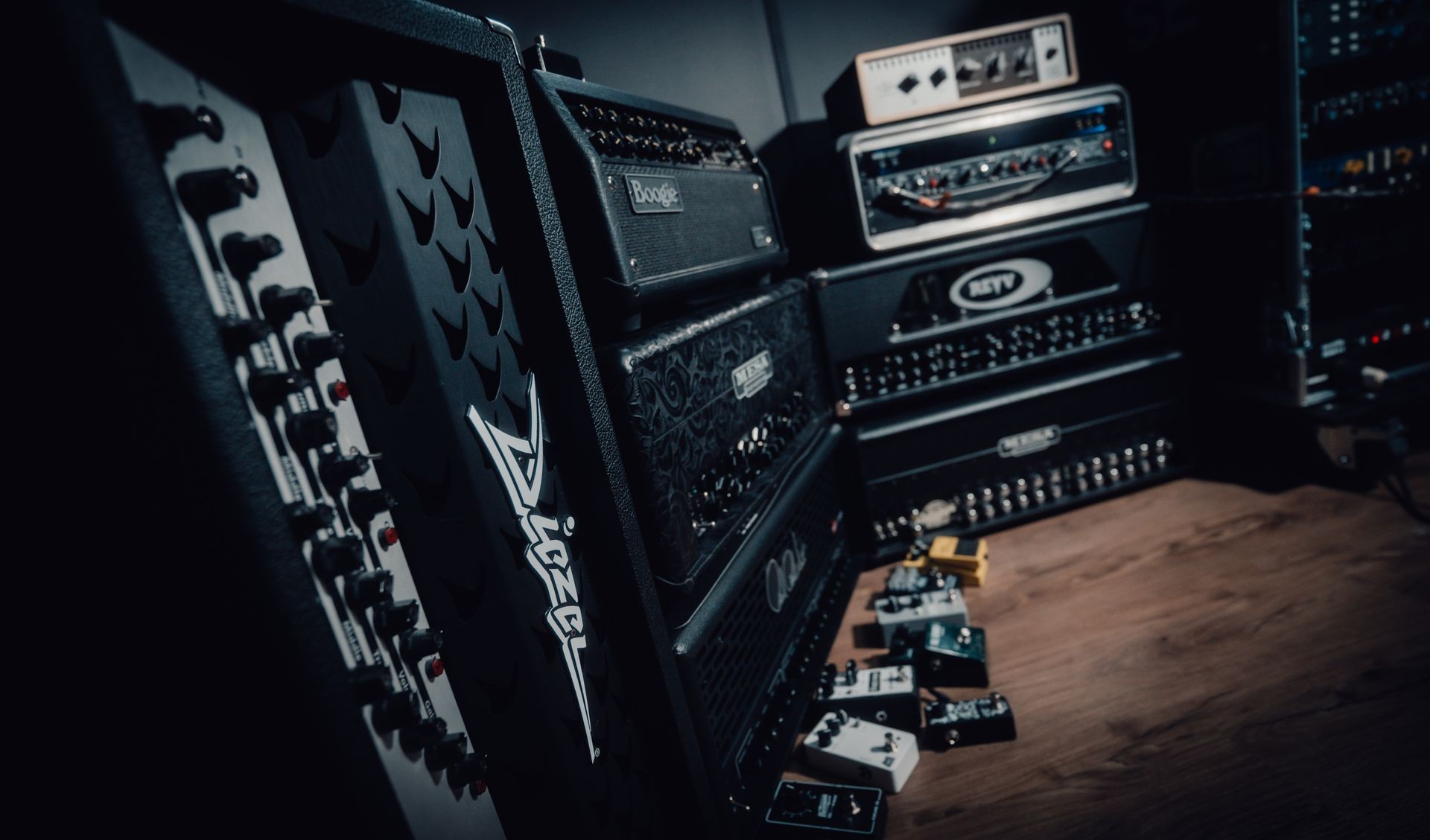 Stack of guitar amplifiers and pedals on a wooden floor in a dimly lit music studio.